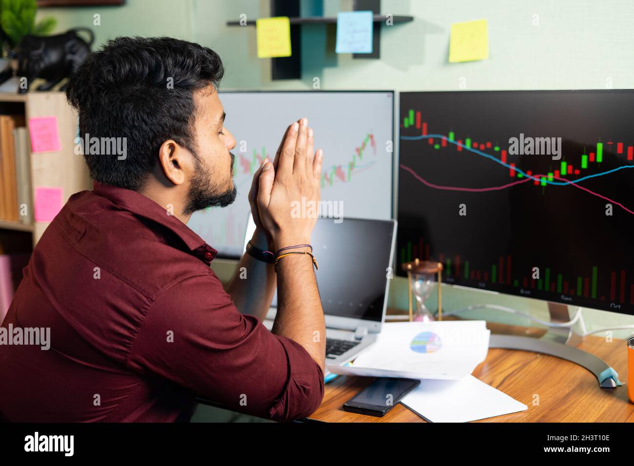 Worried trader praying god to make profit or stock market to go up infront of charts on computer screan - Concept of risk in crypto trading and Stock Photo
