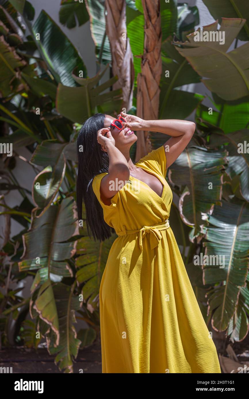 Vertical image of a Latina woman in a yellow dress wearing sunglasses and holding them with both hands looking up against a background of green plants Stock Photo