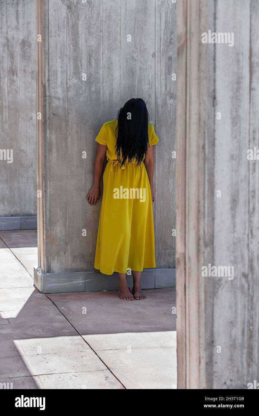 Vertical image of an adult woman with a yellow dress with long black hair covering her face barefoot leaning on a wall or column at daytime. Stock Photo