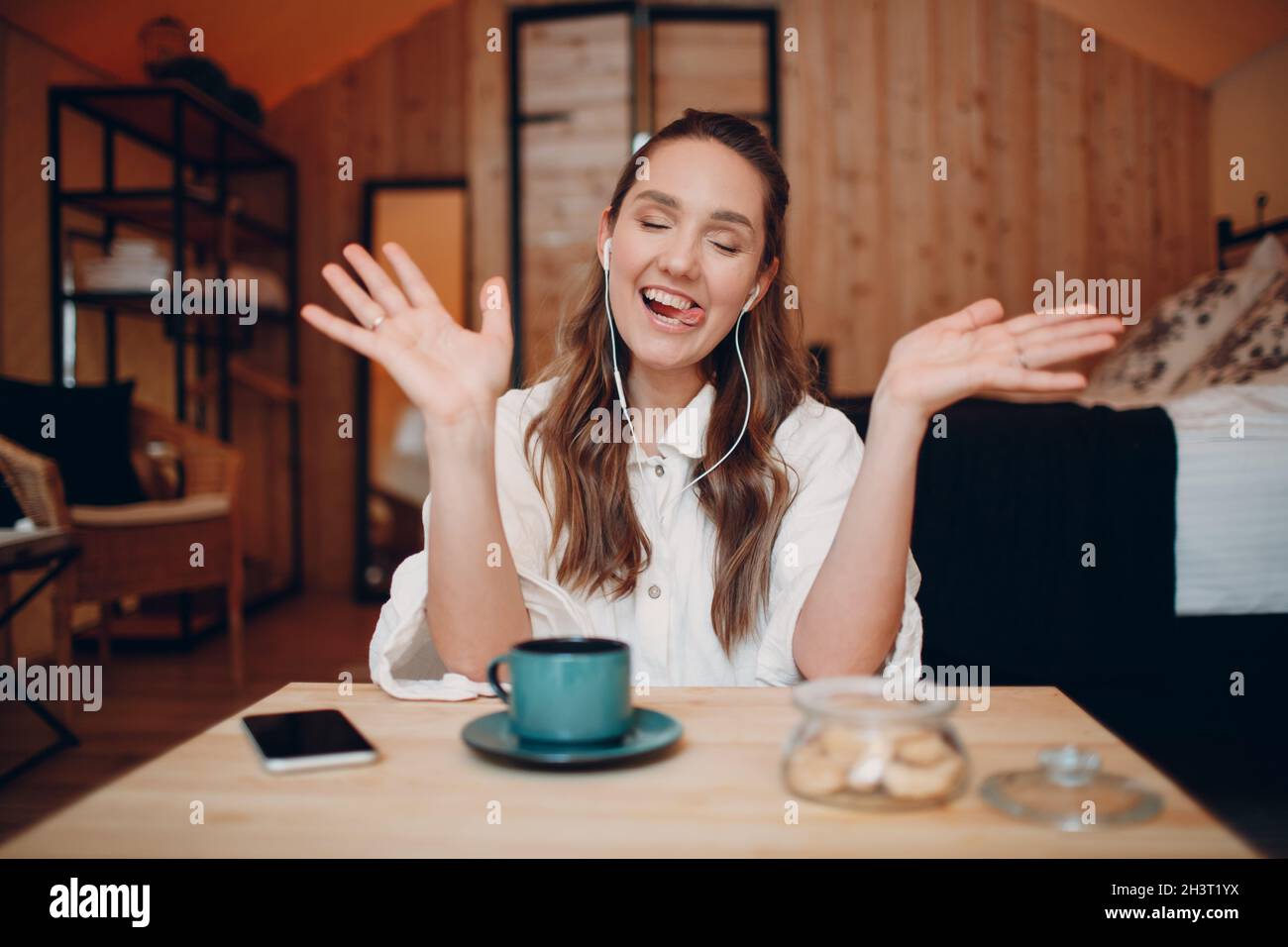 Smiling young woman with headset sitting at table at home behind computer laptop and talking on video call. Female with cup of t Stock Photo