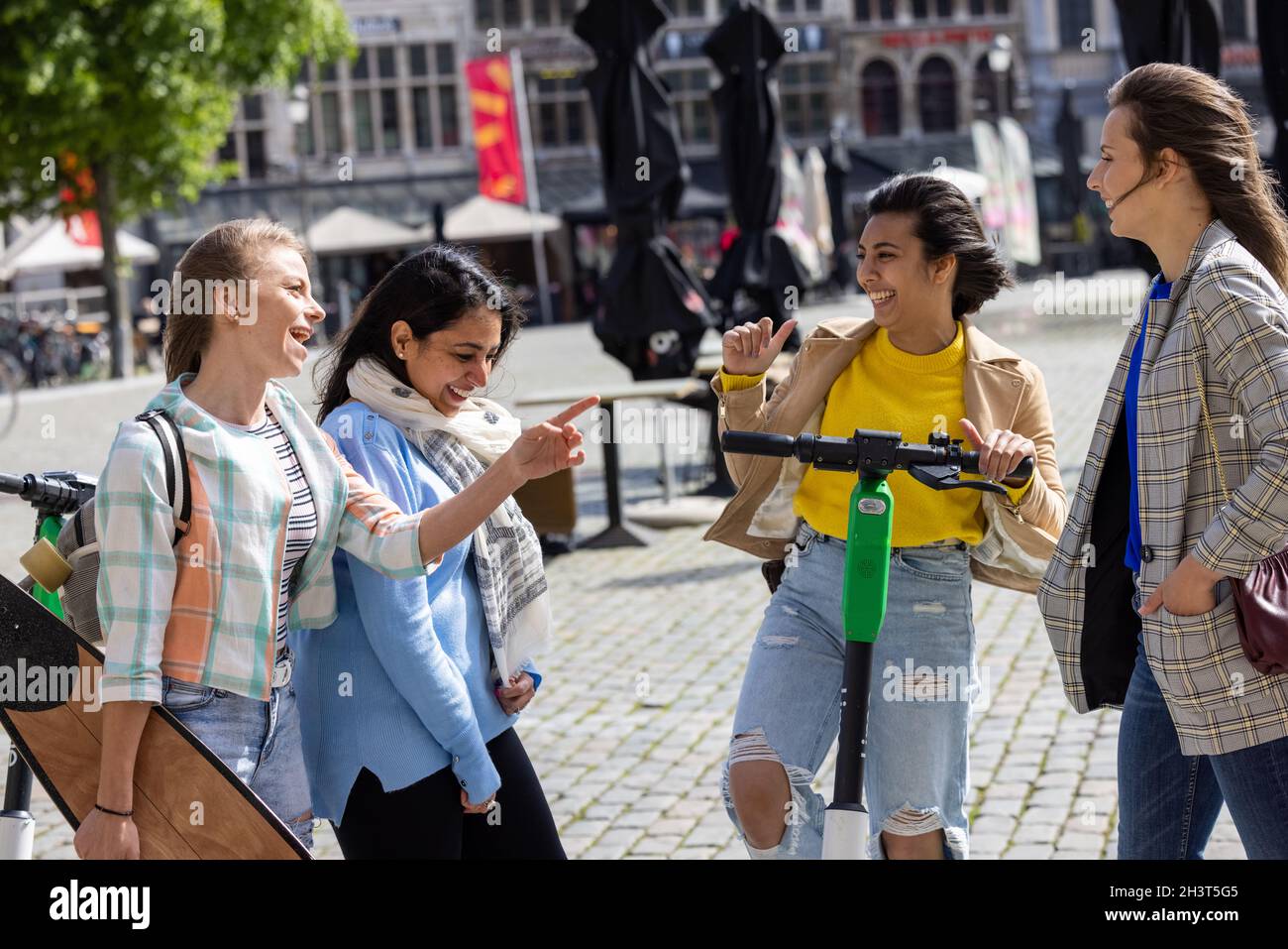 Lifestyle portrait of a diverse multiethnic group of four young girl friends on the electro scooter and skateboard having fun in Stock Photo