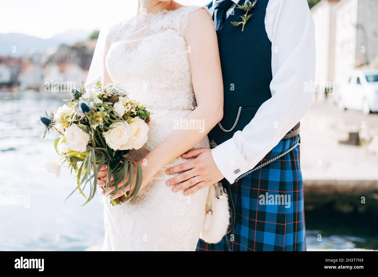 A bride in a wedding dress with bridal bouquet and a groom in a Scottish national dress stand hugging on the pier Stock Photo