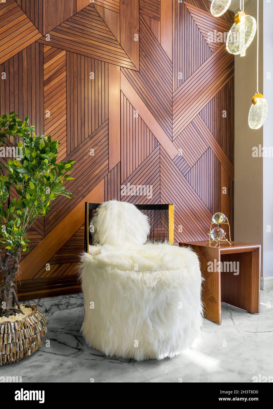 Modern white feather armless chair, small wooden modern table, and planter with green bushes, in a hall with decorated wood cladding wall, and white marble floor Stock Photo