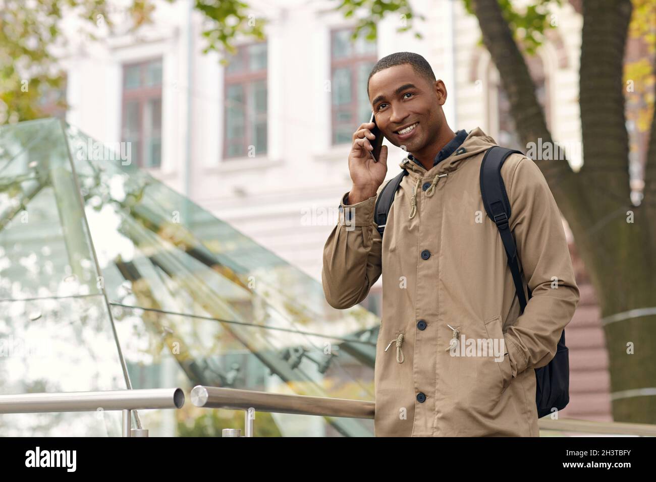 Smiling black male student standing with hand in pocket and talking on mobile phone. Young mulatto man wear parka coat and backpack. Blurred city back Stock Photo