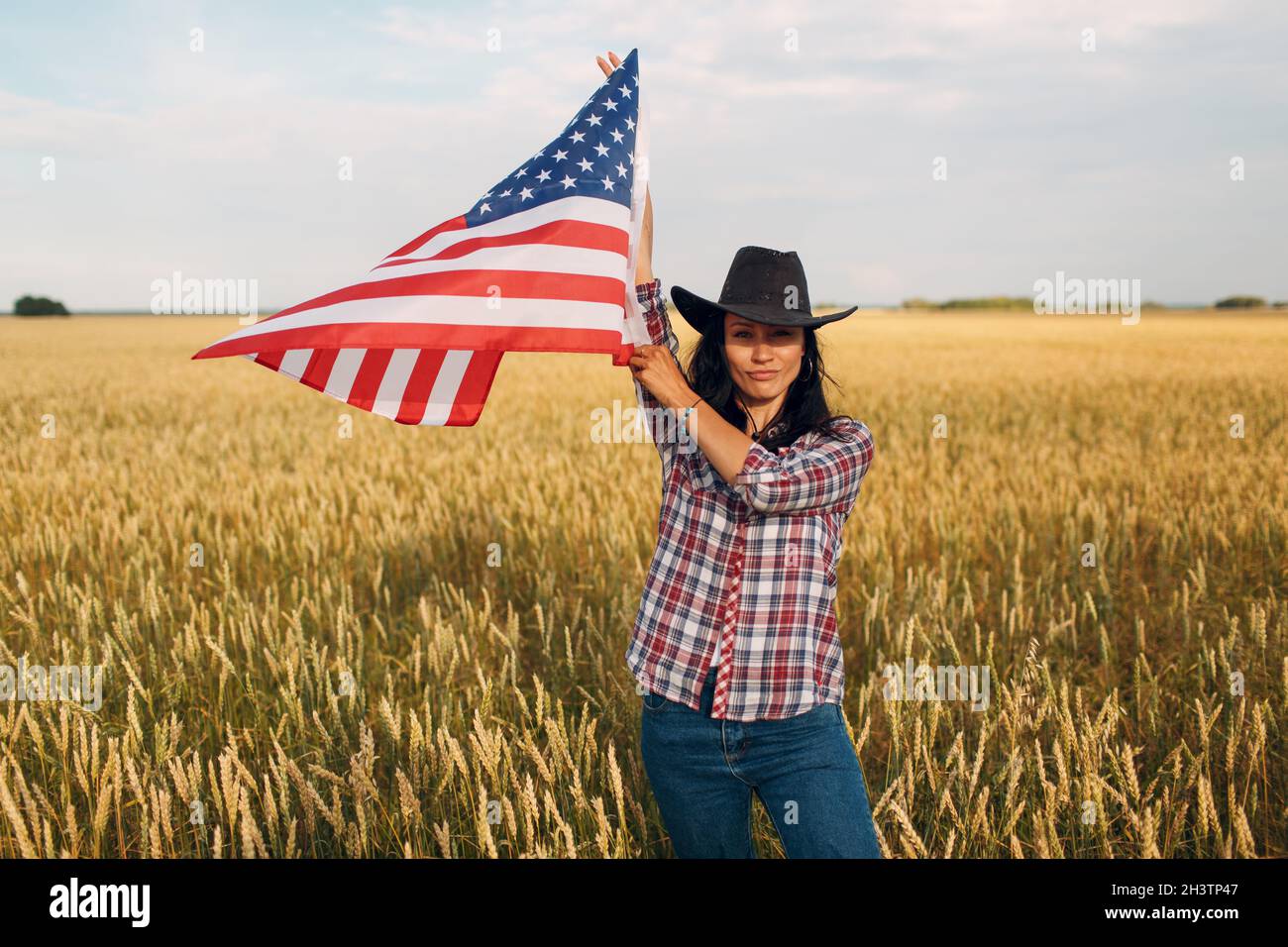 Woman farmer wearing cowboy hat, plaid shirt and jeans waving american flag at wheat field Stock Photo