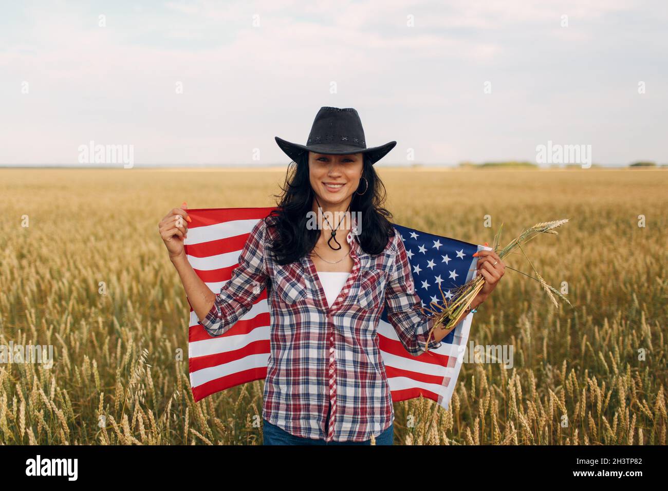 Woman farmer wearing cowboy hat, plaid shirt and jeans with american flag at wheat field Stock Photo