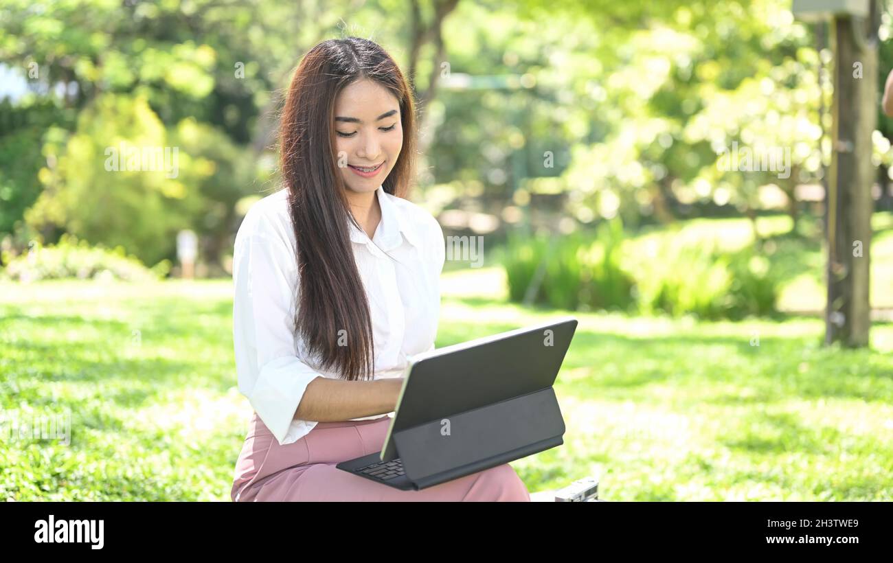 Pleased asian woman sitting in park and using computer tablet Stock ...
