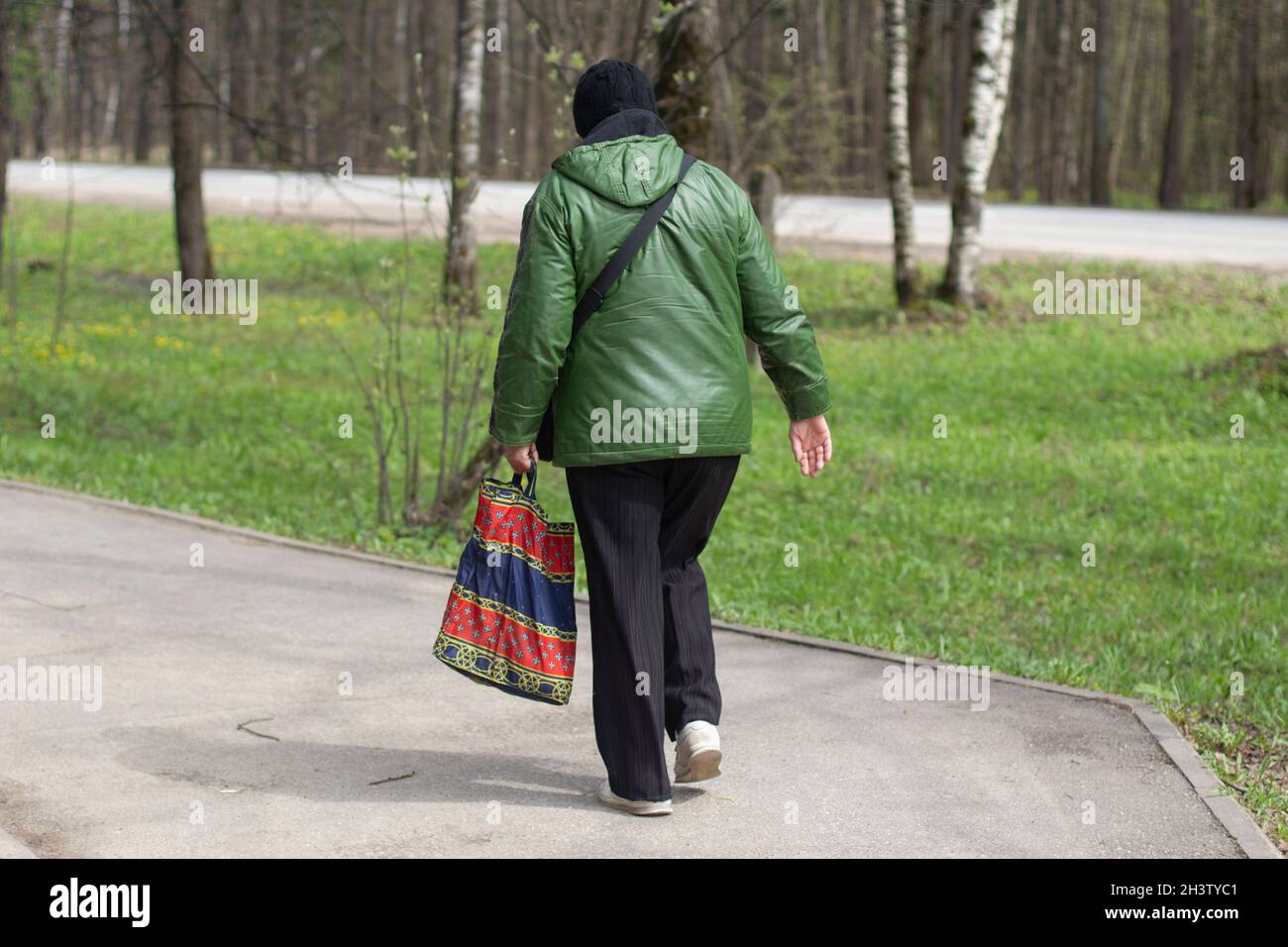 An elderly woman in casual clothes with a bag walks down the street in the park. View from the back. Green jacket, black hat and trousers. Early sprin Stock Photo