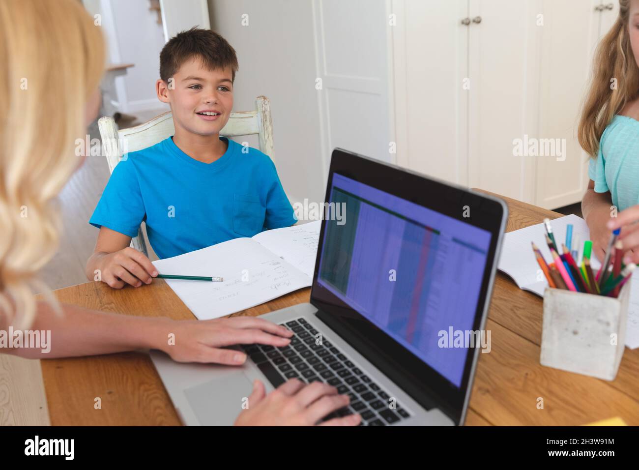 Caucasian mother using laptop and doing homework with her daughter and son smiling in kitchen Stock Photo