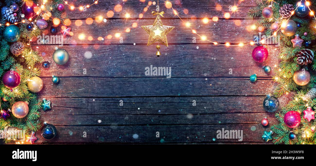 Christmas Decoration - Fir Branches And Baubles Illuminated With String Lights On Dark Wooden Table With Abstract Snowfall Stock Photo