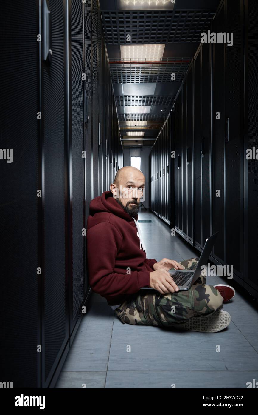 The hacker with a bald head and a beard sits with the computer among servers, the burglar, Storage of data, cloudy services, system breaking, attack Stock Photo