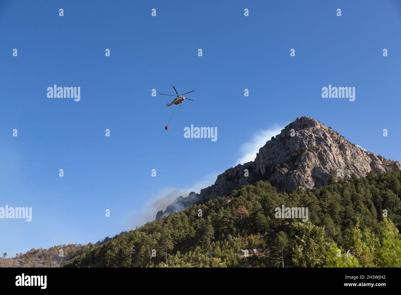 Wildfire helicopter goes to pour water on a fire on the slope of a steep, rocky hill. Stock Photo