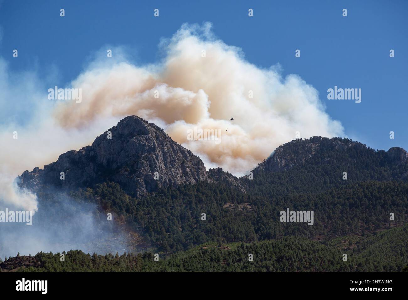 The forest on the slopes of a steep, rocky hill is on fire. Smoke rises into the sky. Wildfire helicopter in the sky. Stock Photo