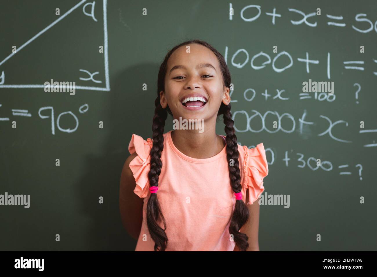 Portrait of laughing mixed race schoolgirl standing in front of chalkboard in classroom maths lesson Stock Photo