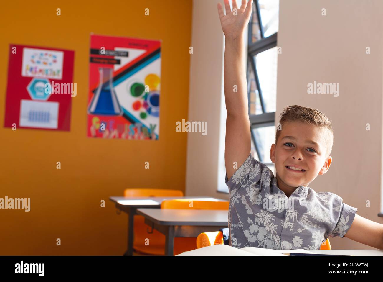 Happy caucasian schoolboy sitting at desk in classroom raising hand ...