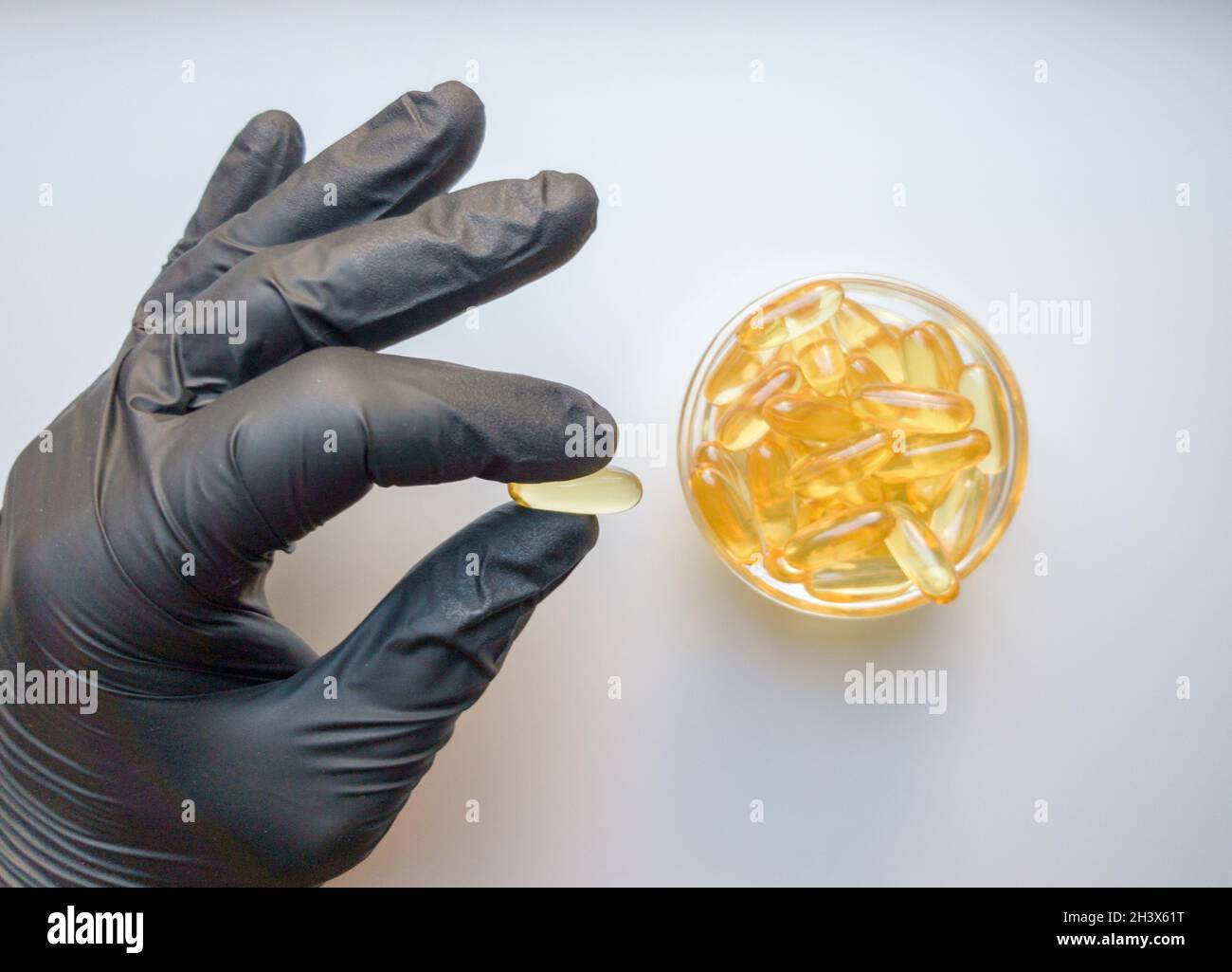 Fish oil capsules in hand in a black medical glove on a white background, the hand takes one tablet from a saucer or plate. Hand Stock Photo