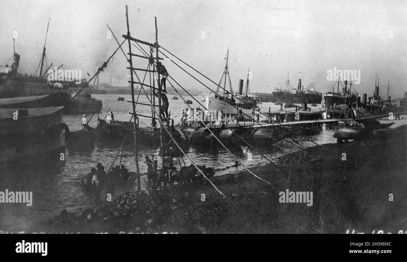 Men on a makeshift ladder in Rangoon harbour, working a pile driver, with ships of various sizes in the background., 1909. Stock Photo