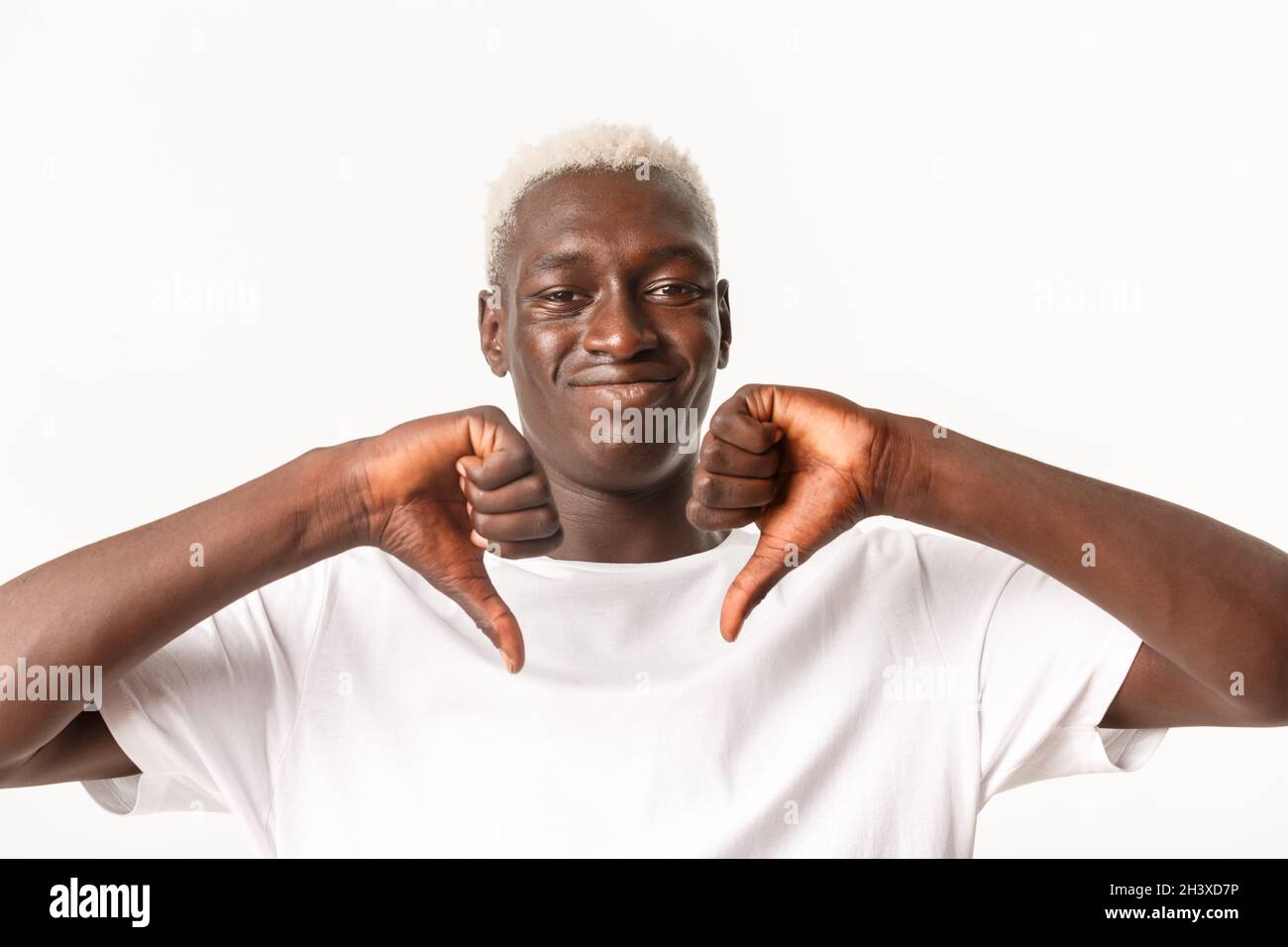 Close-up of unamused and skeptical Black guy with blond hair, showing thumbs-down and smirk with dislike, disapprove bad thing, Stock Photo