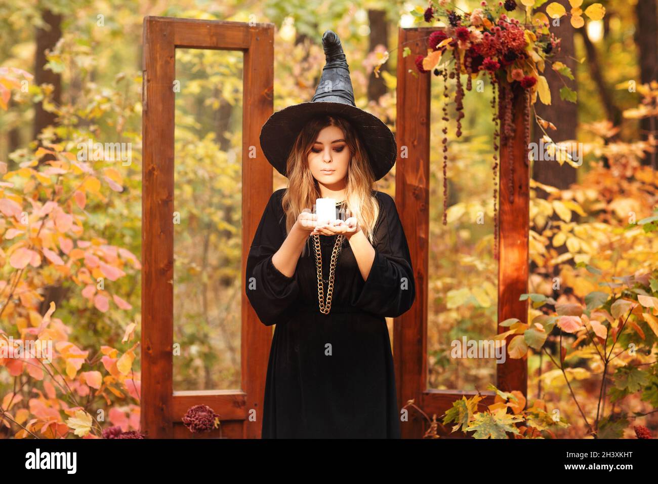 Young woman in Witch costum and hat with candle in hands stands against autumn forest background Stock Photo