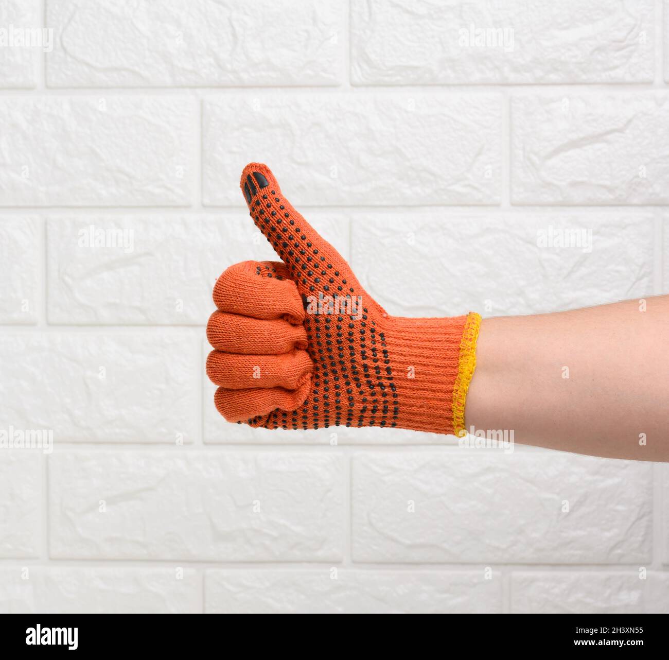 Female hand in an orange work protective glove shows the gesture like on a white brick background Stock Photo