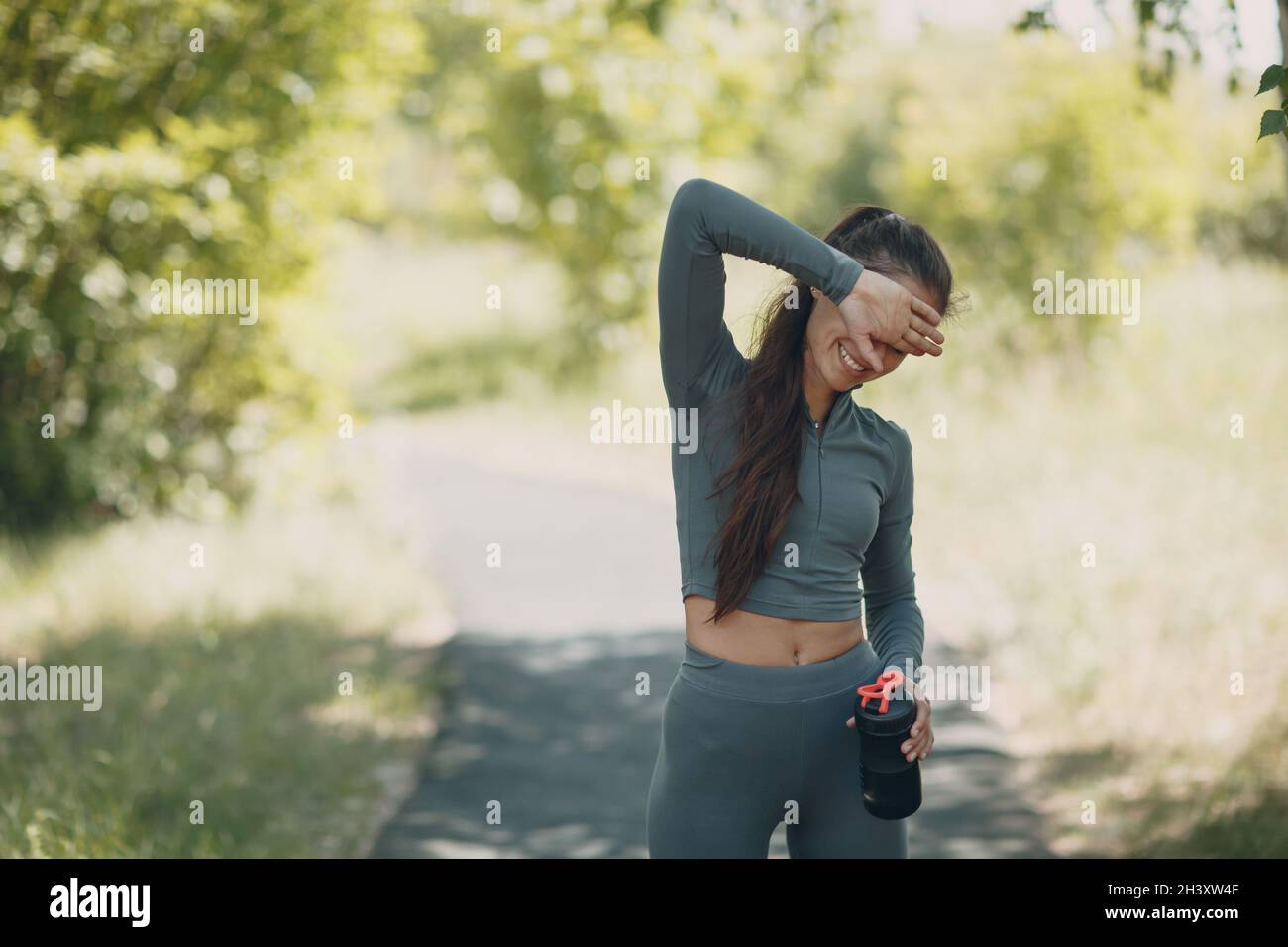 Tired runner woman jogger drinking bottled water after jogging in park outdoor. Wet armpit and deodorant antiperspirant. Stock Photo