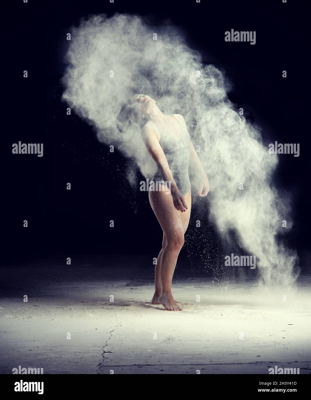 Beautiful caucasian woman in a black bodysuit with a sports figure is dancing in a white cloud of flour on a black background Stock Photo