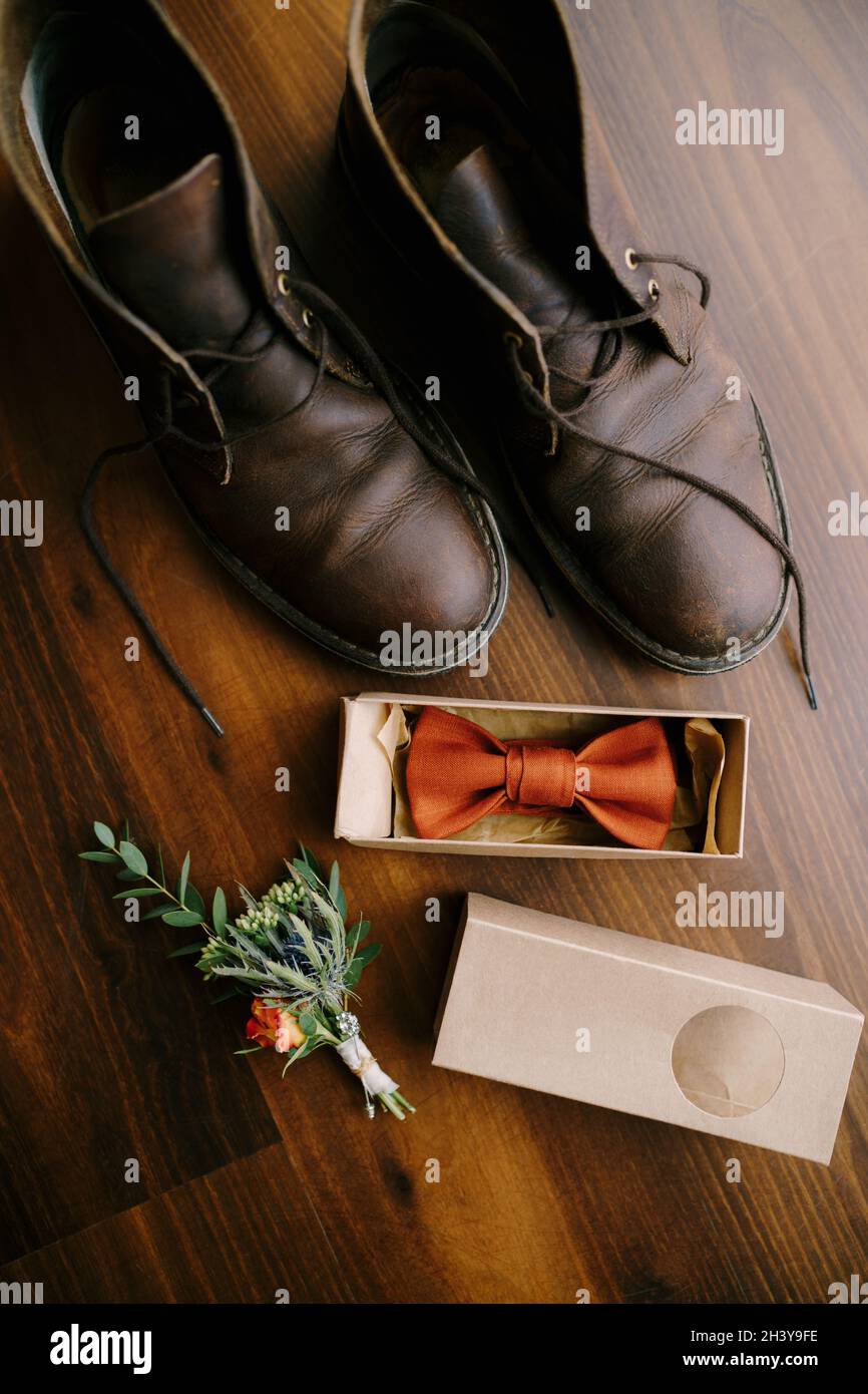 Bow tie in a box for a groom with a boutonniere, and men's boots with untied laces on a brown floor. Stock Photo