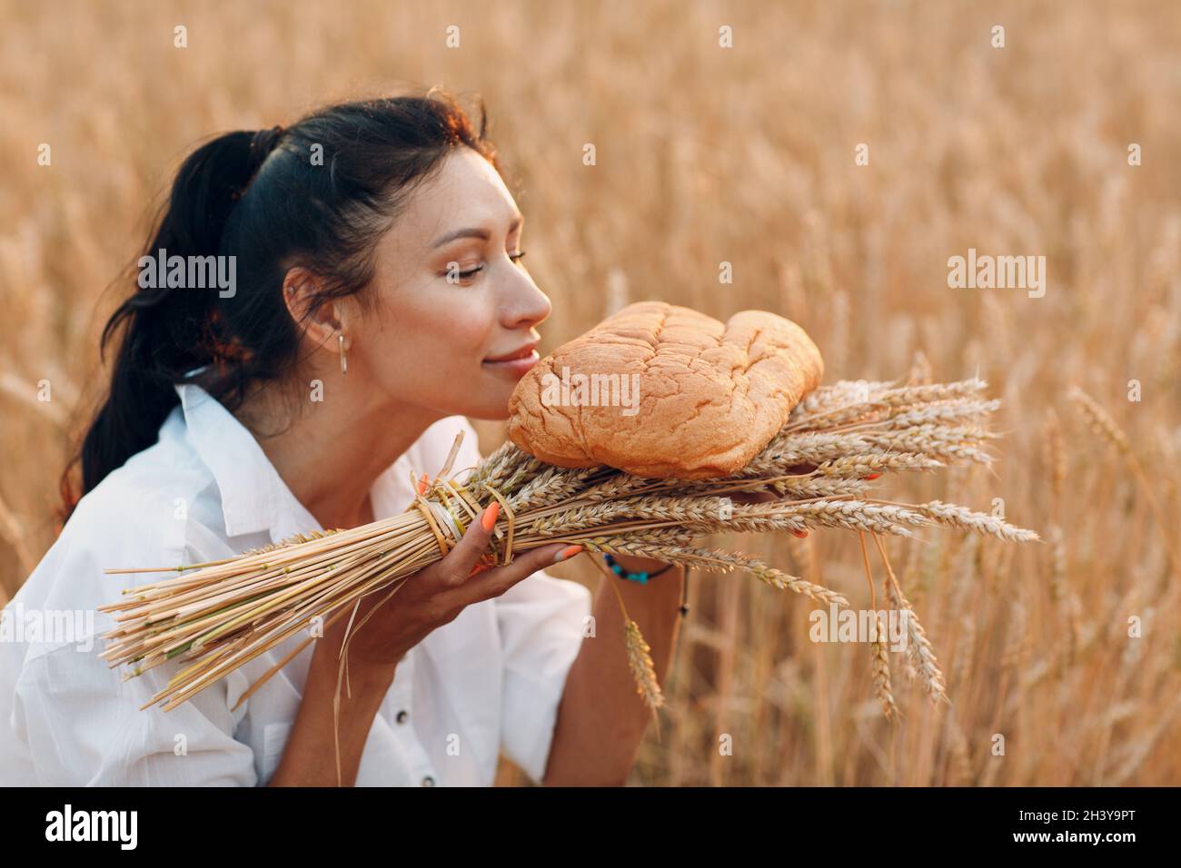 Woman Holding and Smelling Homemade Wheat Bread and Sheaf of Ears In Hands In Wheat Field at Sunset. Stock Photo