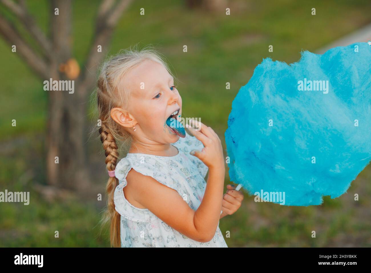 Little blond girl eating cotton candy and shows blue tongue in the park. Stock Photo