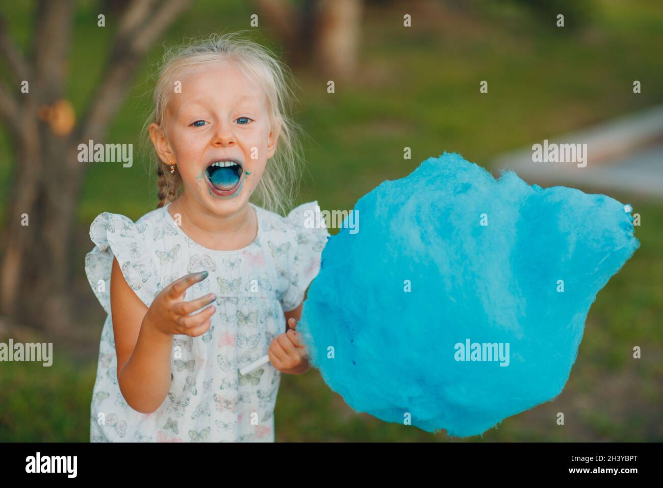 Little blond girl eating cotton candy and shows blue tongue in the park. Stock Photo