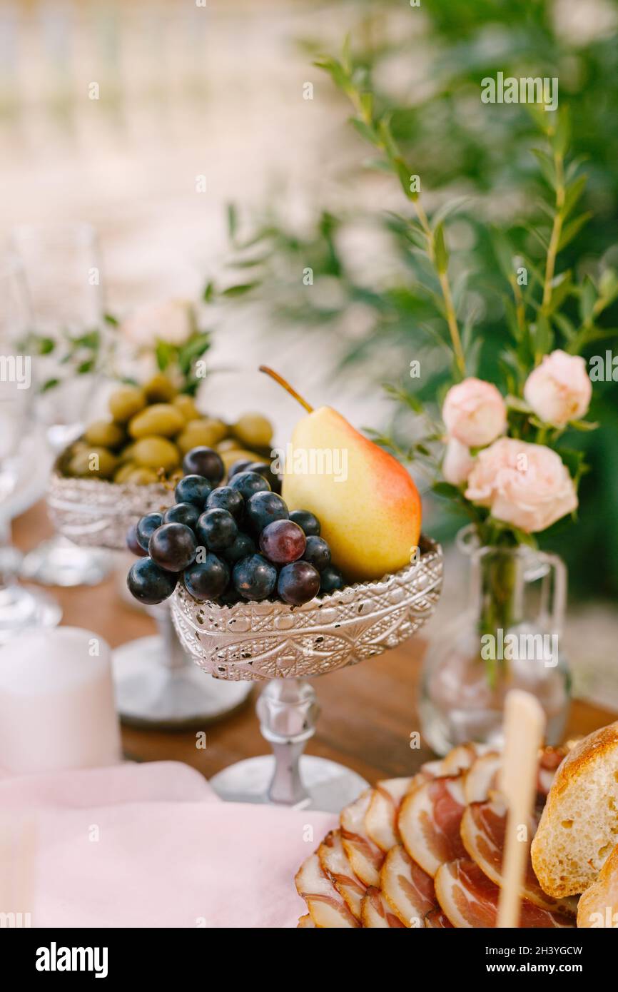 A close-up of a silver-colored bowl with grapes and a pear on the table with ham, a vase with a flower and a bowl of olives. Stock Photo