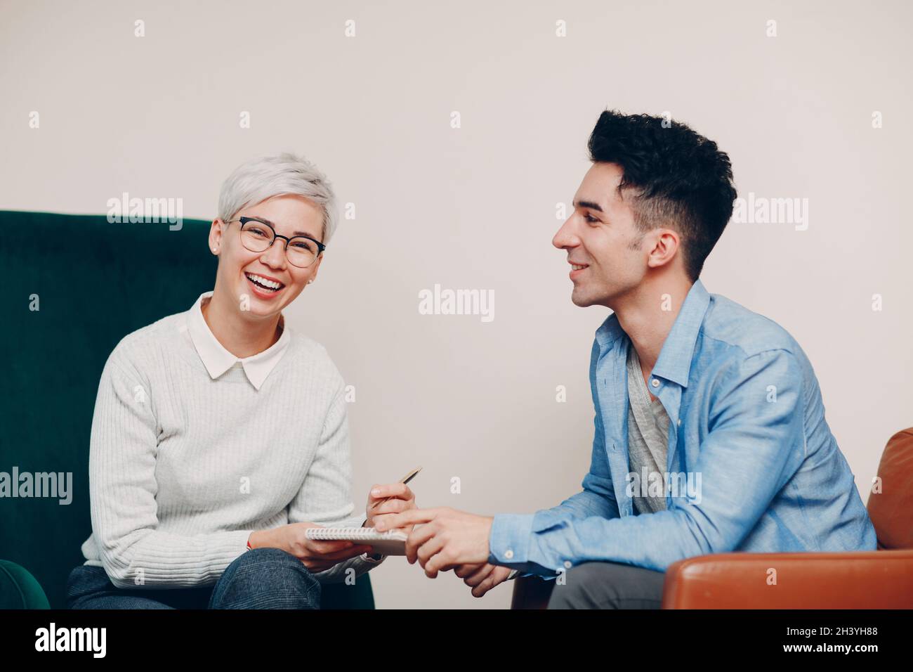 Woman with pen and notebook in hand and man smile sitting on chair together. Journalism, interview and HR recruitment survey con Stock Photo