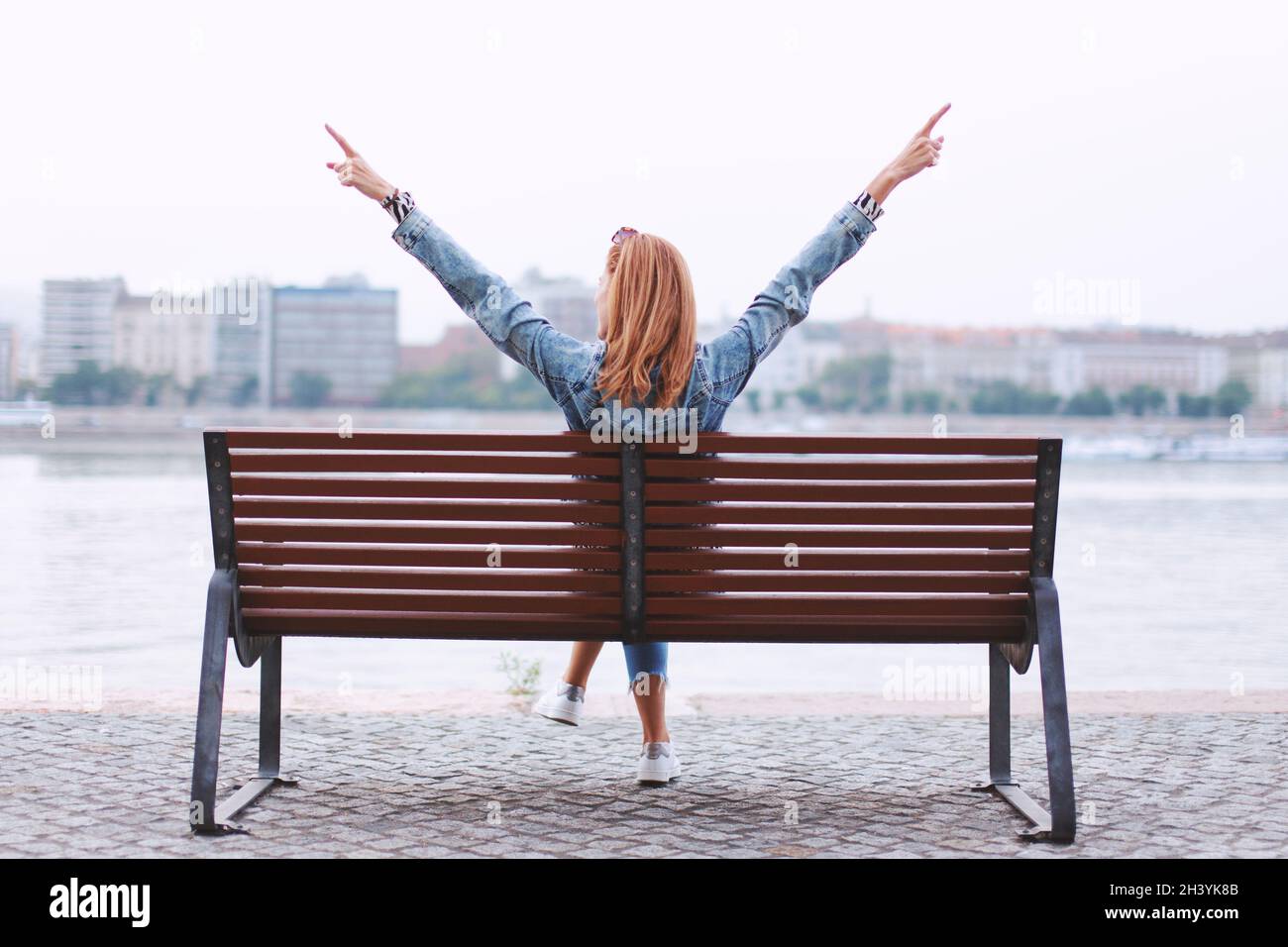Young redhead woman arms raised on bench at riverbank back view Stock Photo