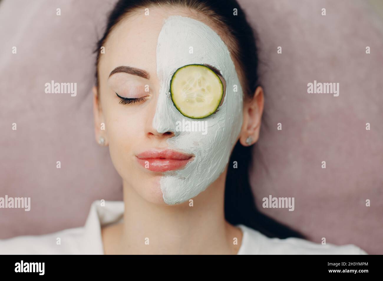 Young woman applying mask of clay on face with cucumber on eyes in beauty spa Stock Photo