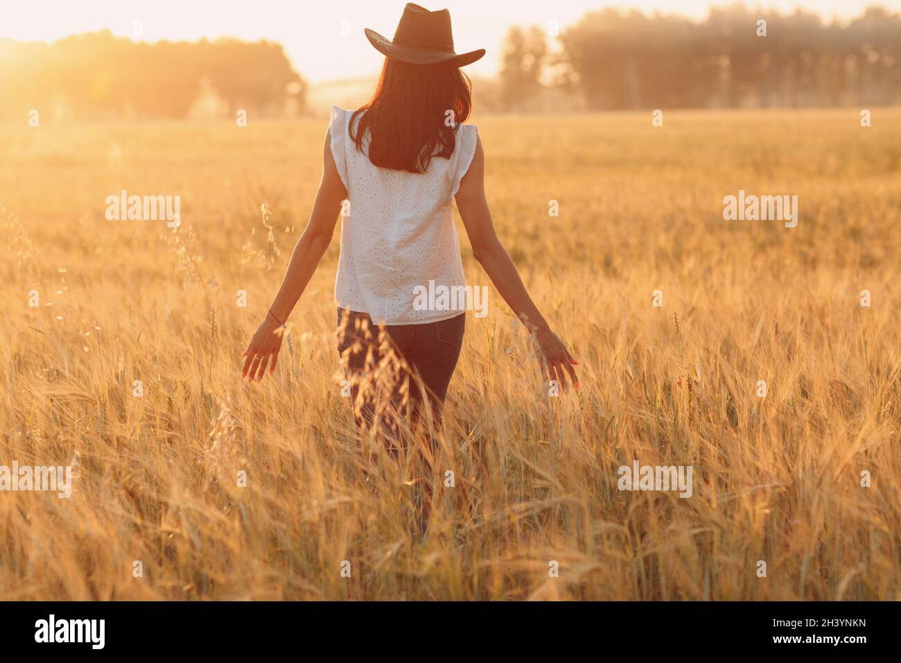 Woman farmer in cowboy hat walking with hands on ears at agricultural wheat field on sunset Stock Photo