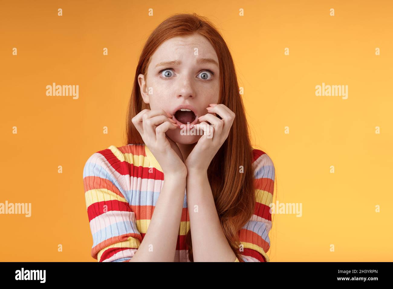 Timid insecure young ginger girl blue eyes feeling scared gasping shocked frightened trembling fear hold fingers mouth frowning Stock Photo