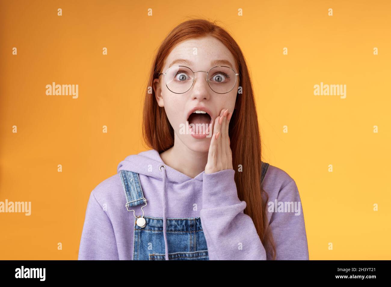 Omg no way. Attractive shocked wondered redhead amused hipster woman modern teenager drop jaw gasping wide eyes surprised standi Stock Photo