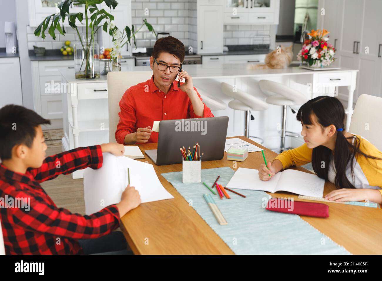 Asian father working on laptop at table with son and daughter doing school work Stock Photo