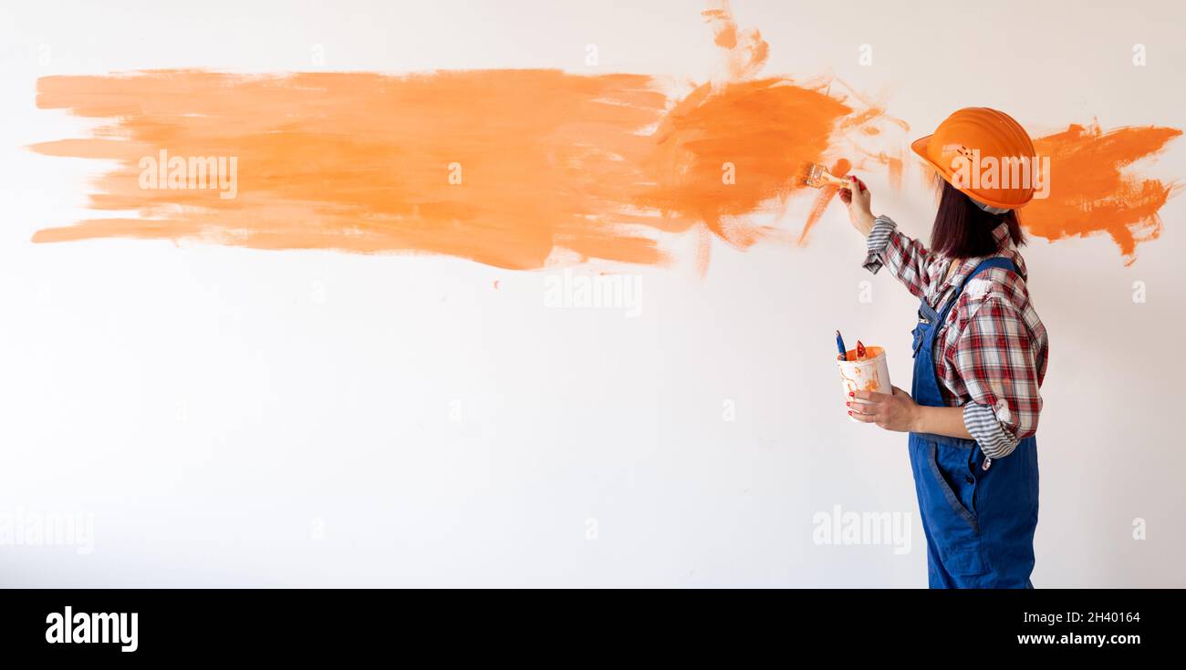 A woman in overalls and a helmet paints the wall in the room with orange paint with a brush. The concept of home renovation and painting the walls. Stock Photo