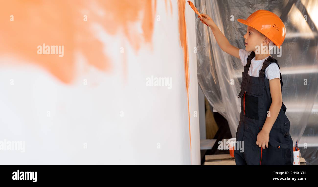 The boy paints the wall with a brush with orange paint. A child dressed in a helmet and overalls makes house repairs indoors. renovation concept Stock Photo