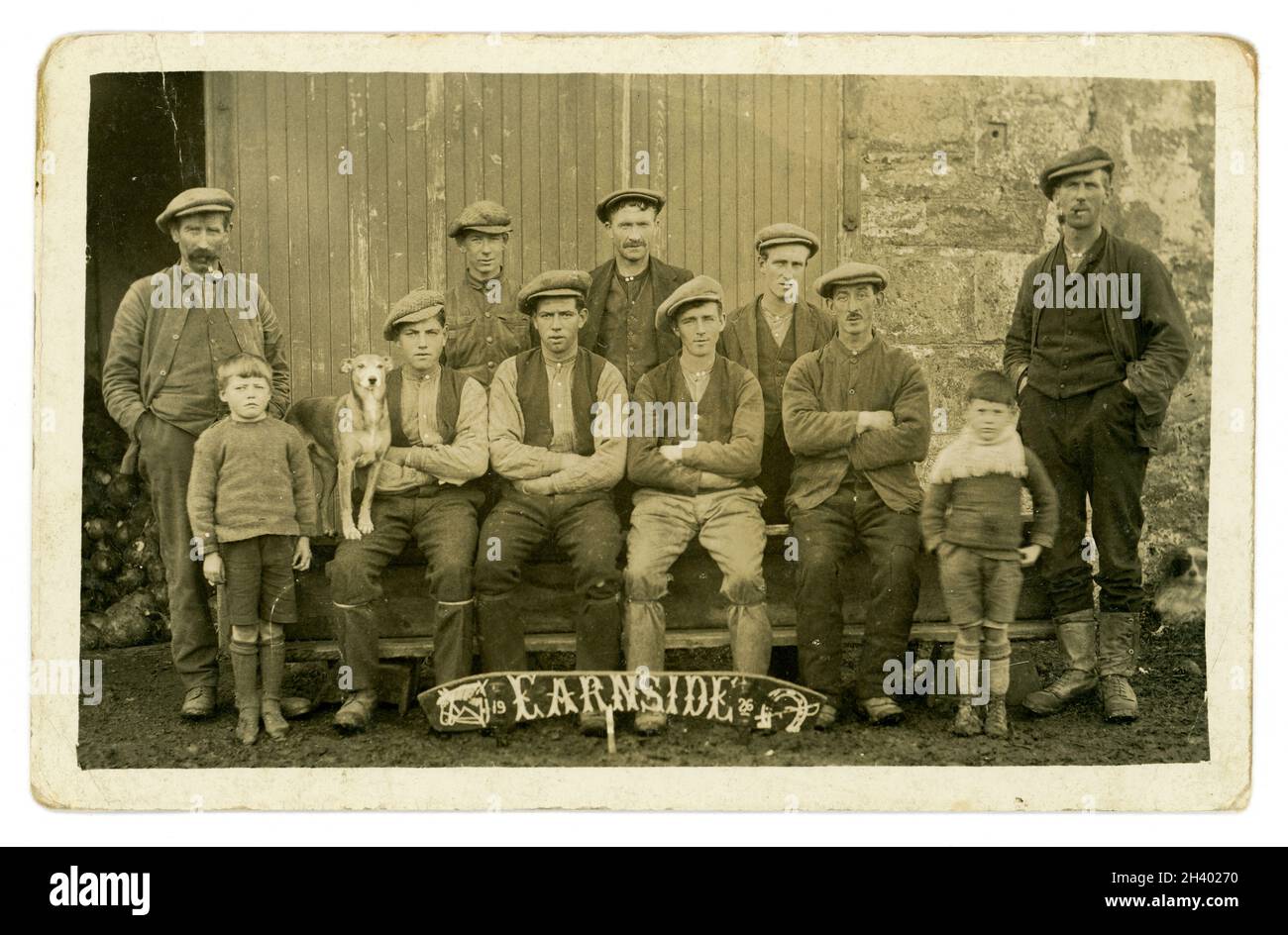 Original early 1920's postcard of stable hands or farm workers - ploughmen, some with their small sons, wearing working clothes - flat caps, waistcoats and trousers tied below knee, boots. Posing for a group photo outside a barn building or stables with a dog. They appear to be working for a large landowner. They could possibly be working with Clydesdale draught horses for ploughing  At their feet is a wooden sign dated 1926, which also has Earnside, a horseshoe, whip and horse drawn on it. Possibly a celebration or anniversary. Located near the river Earn, Perth, Central Scotland, U.K. Stock Photo
