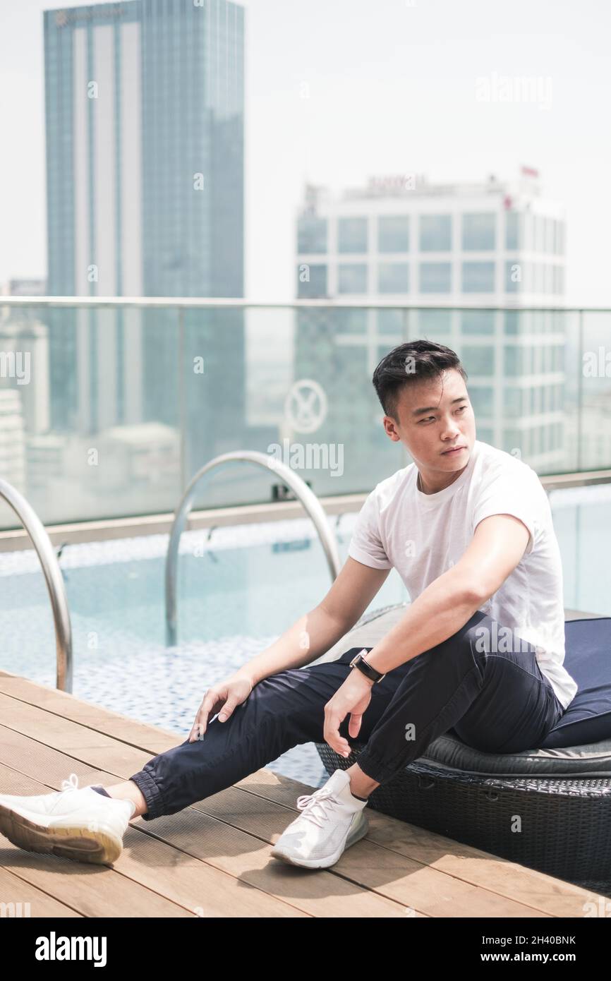 Handsome young Asian man sitting on the rooftop near the swimming pool. Chinese guy, millennials life, lifestyle in the big city, Modern Asia Stock Photo