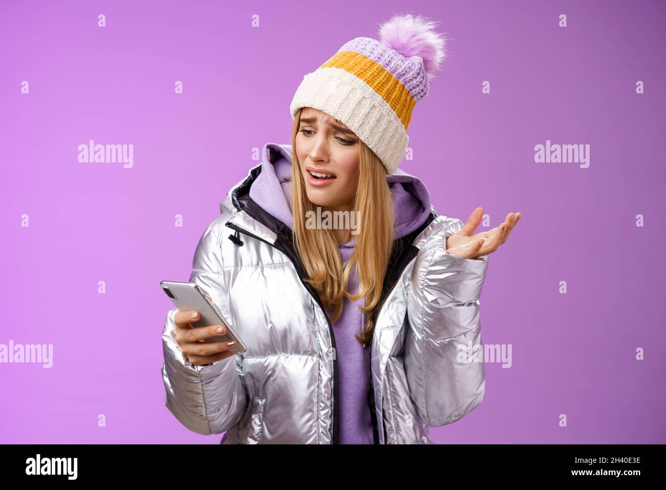 Upset disappointed attractive whining gloomy blond girl in silver jacket standing outside hat holding smartphone shrugging raisi Stock Photo
