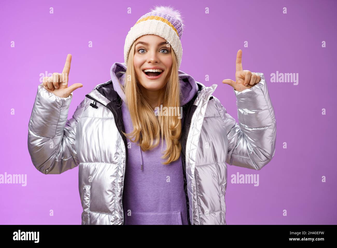 Excited carefree cheerful fair-haired european girl in silver jacket winter hat raising hands pointing up have excellent idea sm Stock Photo
