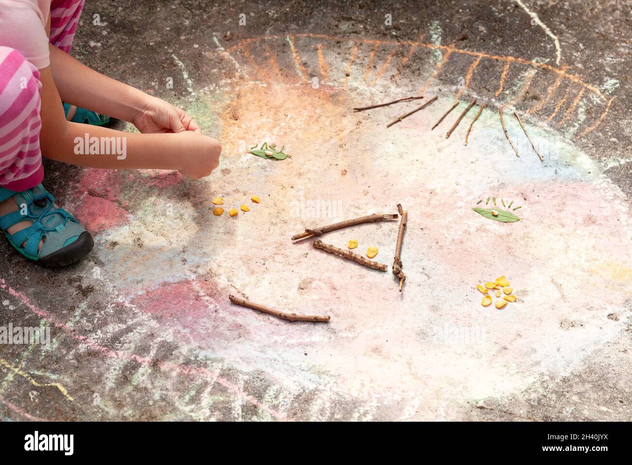 Anonymous young elementary school age child, little girl playing outside, outdoors creating a face shape from sticks, leaves and chalk. Children art t Stock Photo