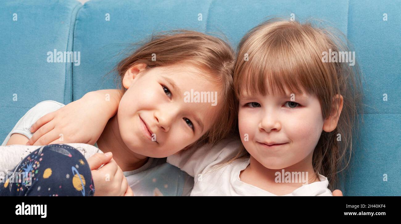 Two young cheerful elementary primary school age girls smiling, sisters, siblings hugging, sitting together on a sofa, portrait, face closeup. Family Stock Photo