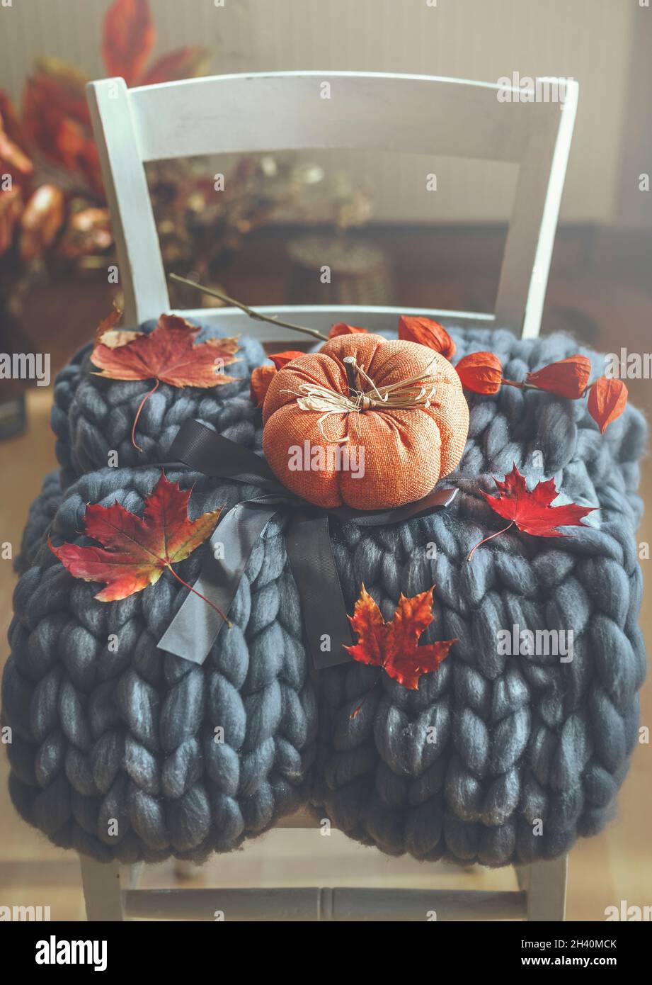 Grey wool blanket with orange pumpkin and autumn leaves on white chair with interior background. Cozy fall time at home with warm clothes and decorati Stock Photo