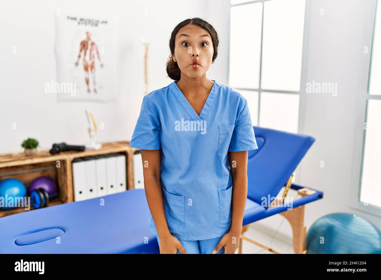 Beautiful hispanic physiotherapist woman at pain recovery clinic making fish face with lips, crazy and comical gesture. funny expression. Stock Photo