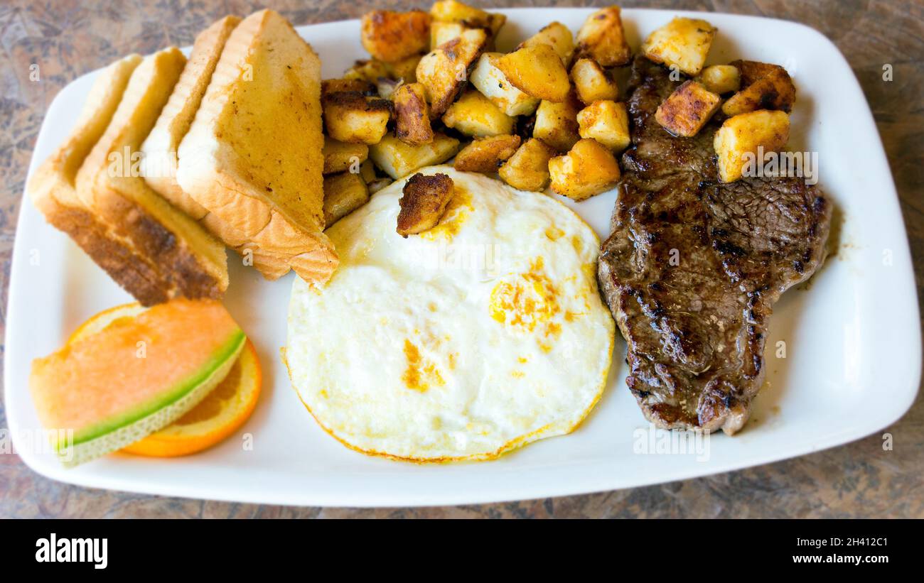 High protein breakfast plate: Sirloin stake and fried eggs. Sides dishes and garnish include potatoes, cantaloupe,  orange slice with toasts socked in Stock Photo