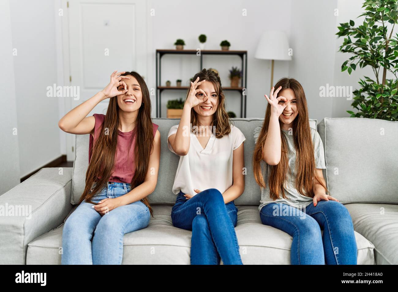 Group of three hispanic girls sitting on the sofa at home doing ok gesture with hand smiling, eye looking through fingers with happy face. Stock Photo